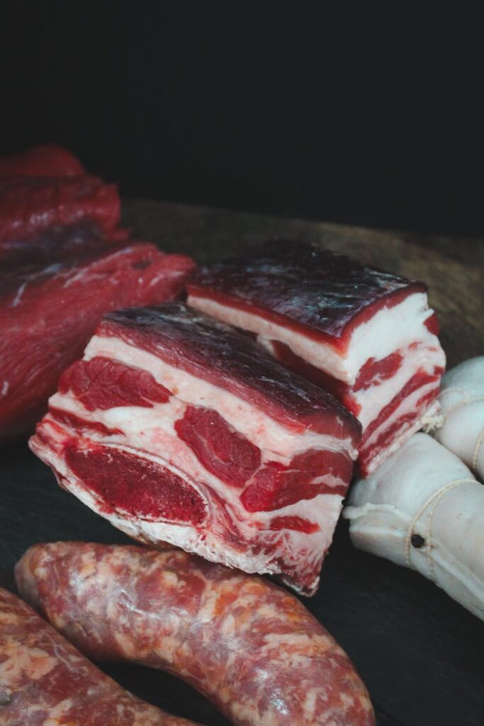 A diverse selection of raw meats including ribs and sausages on a dark surface.
