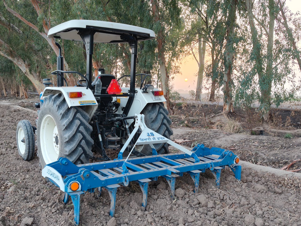 A tractor on a farm in HR, India, working the soil during a vibrant sunset.